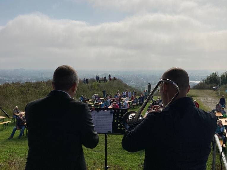 Zwei von hinten zu sehende Musiker auf einem Berggipfel vor Publikum auf Bierzeltgarnituren